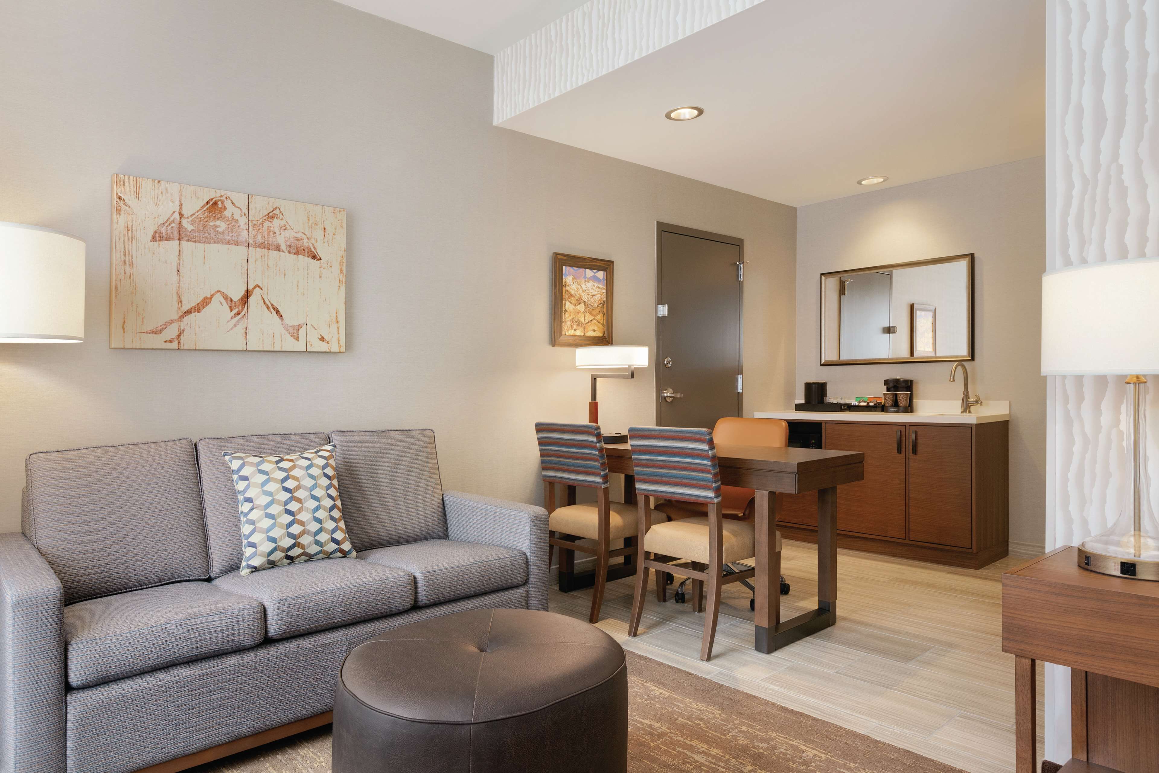 Ein modernes Wohnzimmer mit einem grauen Sofa, einem runden Hocker und einem Essbereich mit einem Holztisch und gestreiften Stühlen.