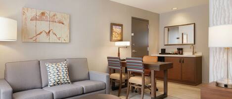 A modern living room with a grey sofa, a round ottoman, and a dining area with a wooden table and striped chairs.