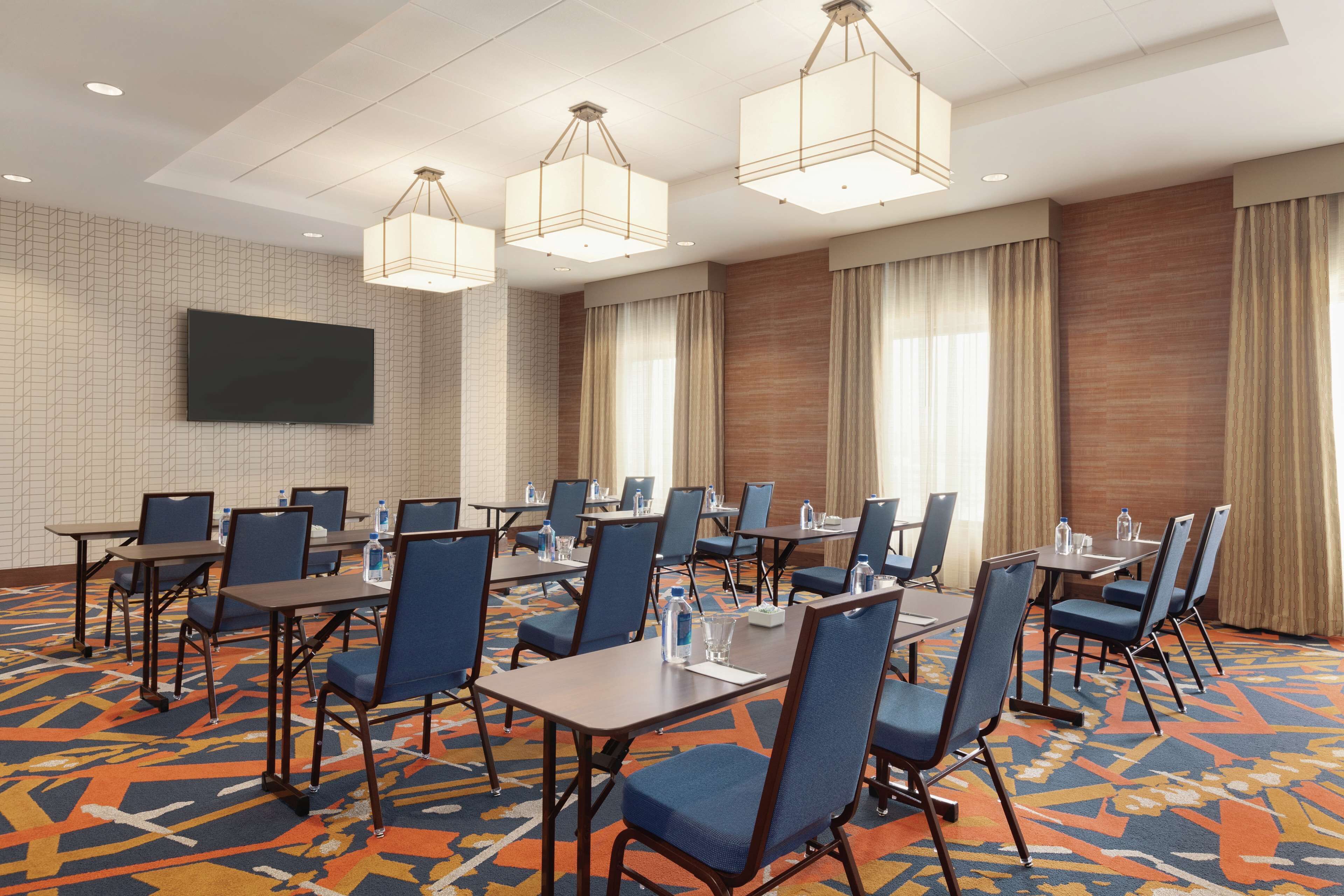A conference room with multiple tables and chairs, a flat-screen TV mounted on the wall, and a patterned carpet.