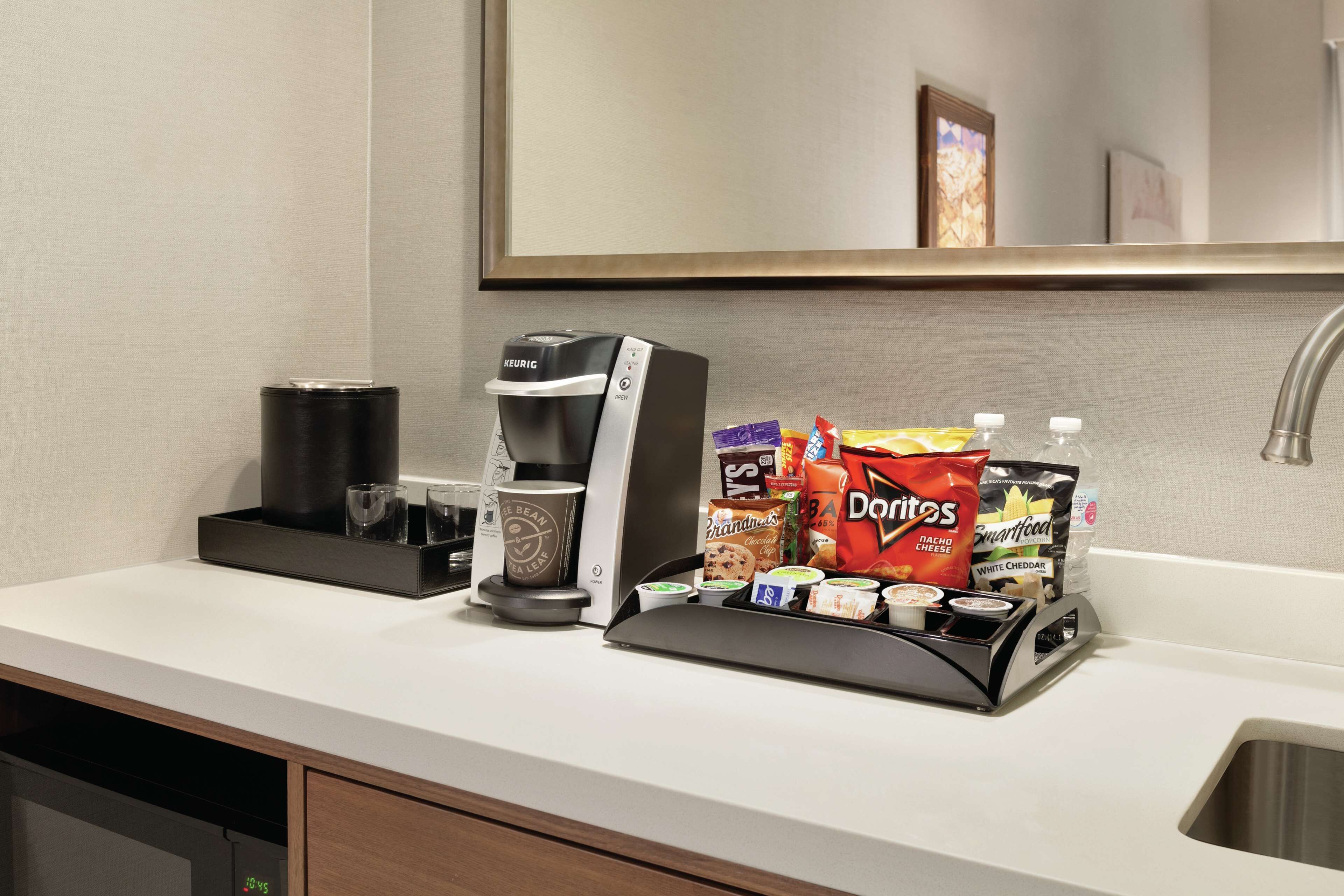 A hotel kitchenette with a Keurig coffee maker, a coffee pot, and a snack station with chips and crackers.