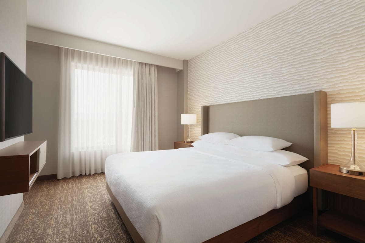 A hotel room with a large bed, a flat-screen TV, a bedside table with a lamp, and a window with curtains.