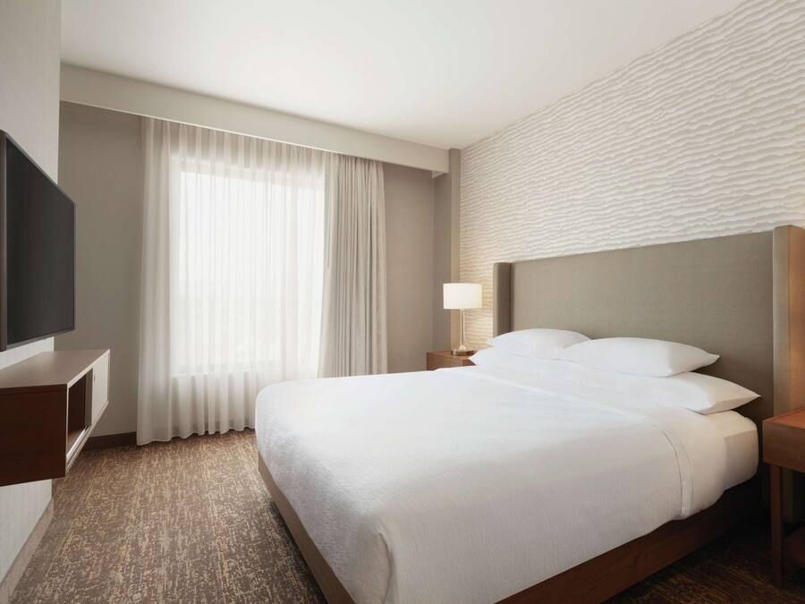 A hotel room with a large bed, a flat-screen TV, a bedside table with a lamp, and a window with curtains.