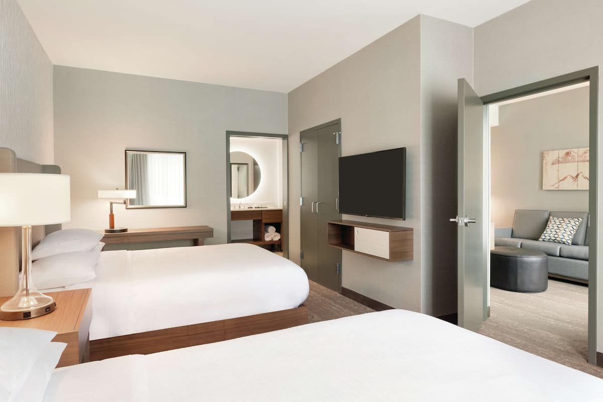 A hotel room with two beds, a TV, a mirror, a desk with a lamp, and a sofa in the adjacent living area.