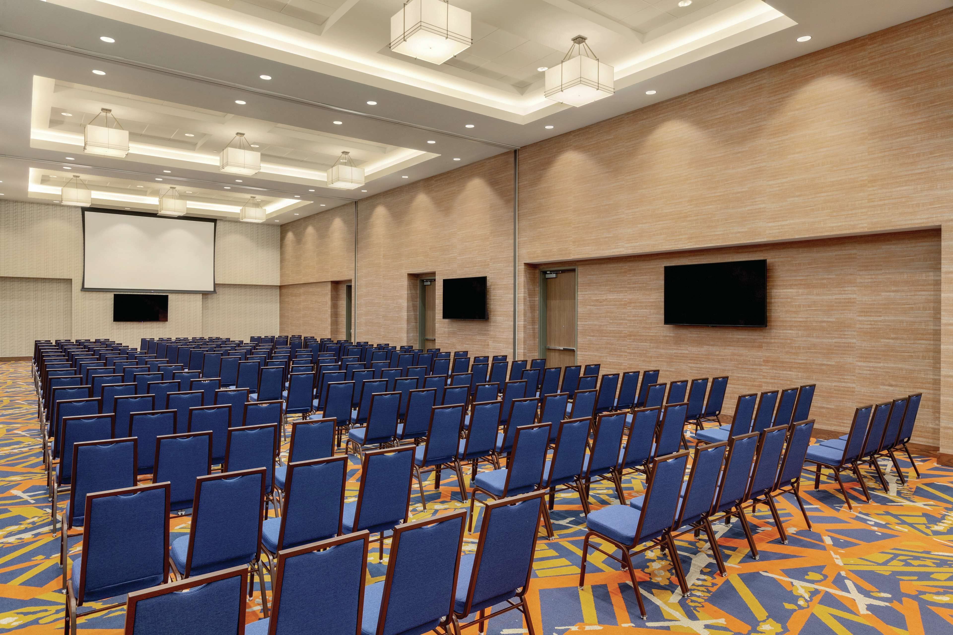 A conference room with rows of blue chairs, a large screen, and a patterned carpet.