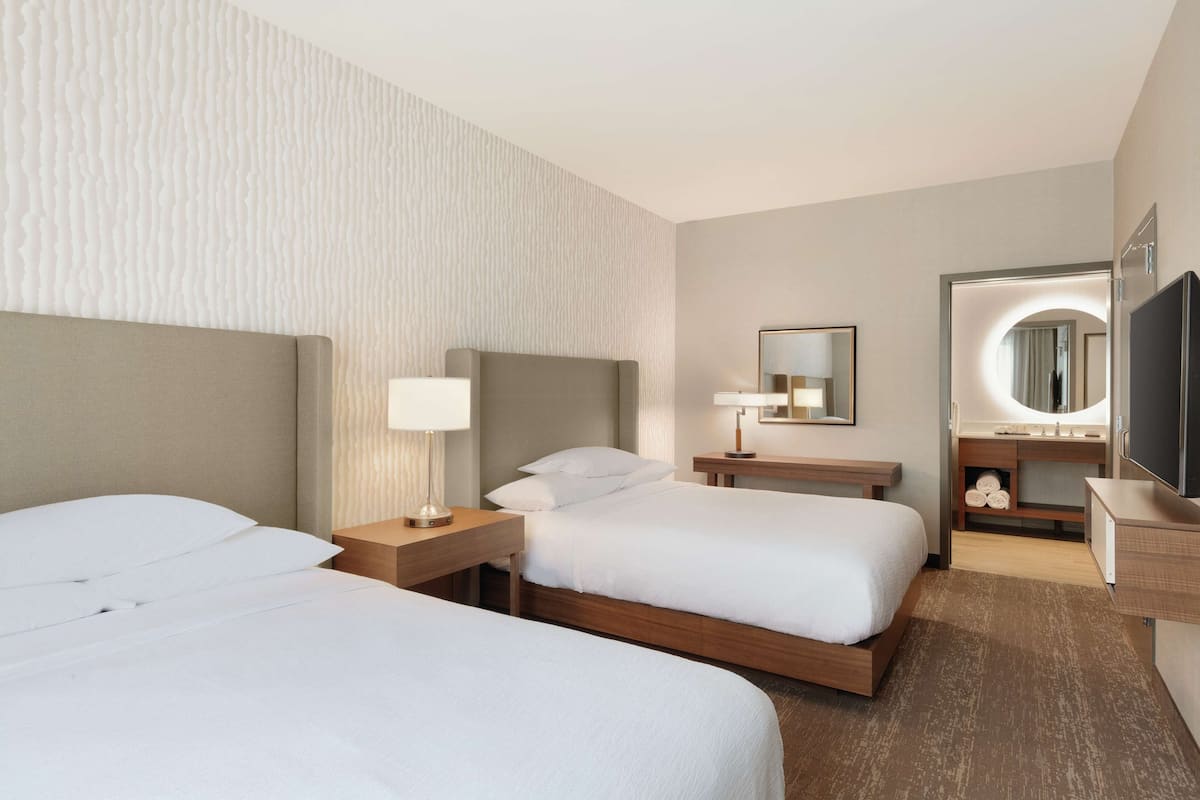 A hotel room with two beds, a vanity with a mirror, and a flat-screen TV.