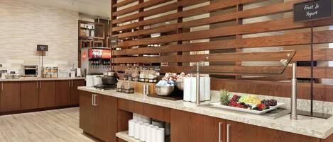 Un espace pour le petit-déjeuner à l’hôtel, avec un buffet de fruits et de yaourts, un réfrigérateur rempli de Coca-Cola et une machine à café.