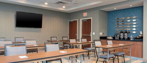 A conference room with multiple tables, chairs, a flat-screen TV, and a kitchen area with appliances.