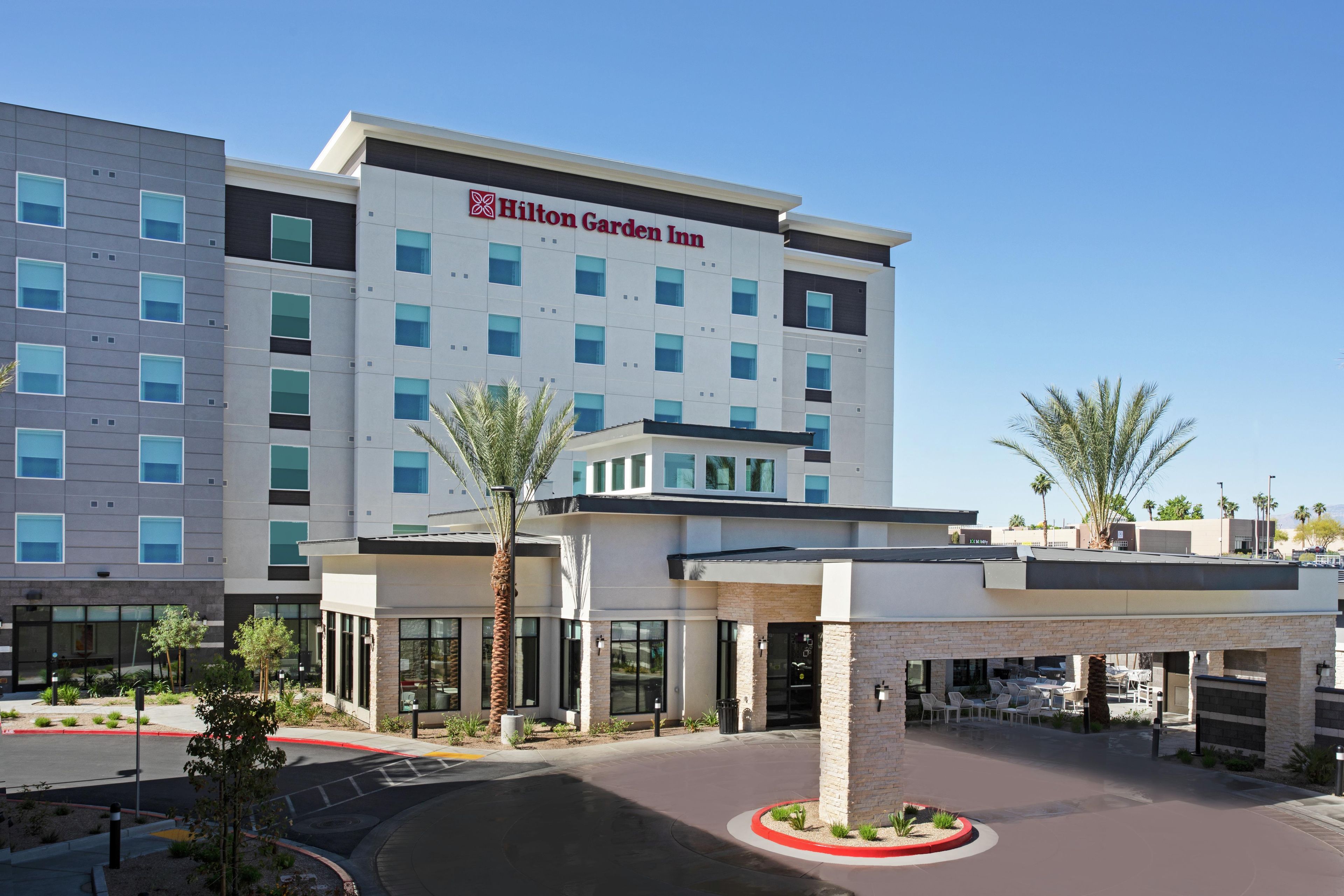 Un Hilton Garden Inn con diseño moderno, que cuenta con un logotipo destacado, un área exterior cubierta con asientos y un estacionamiento.