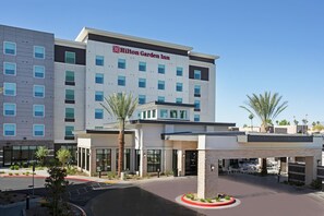 Un Hilton Garden Inn con diseño moderno, que cuenta con un logotipo destacado, un área exterior cubierta con asientos y un estacionamiento.