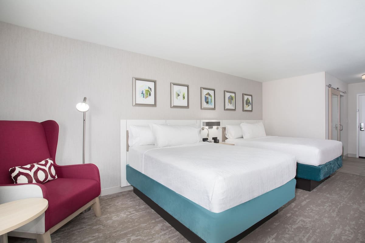 Una habitación de hotel con una cama, un sillón rojo, una mesita y una lámpara de pared.