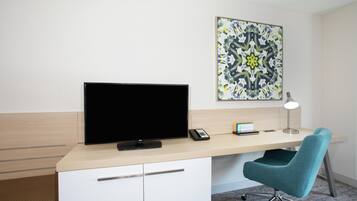 A modern hotel room with a flat-screen TV, a desk with a lamp, a chair, and a decorative wall piece.