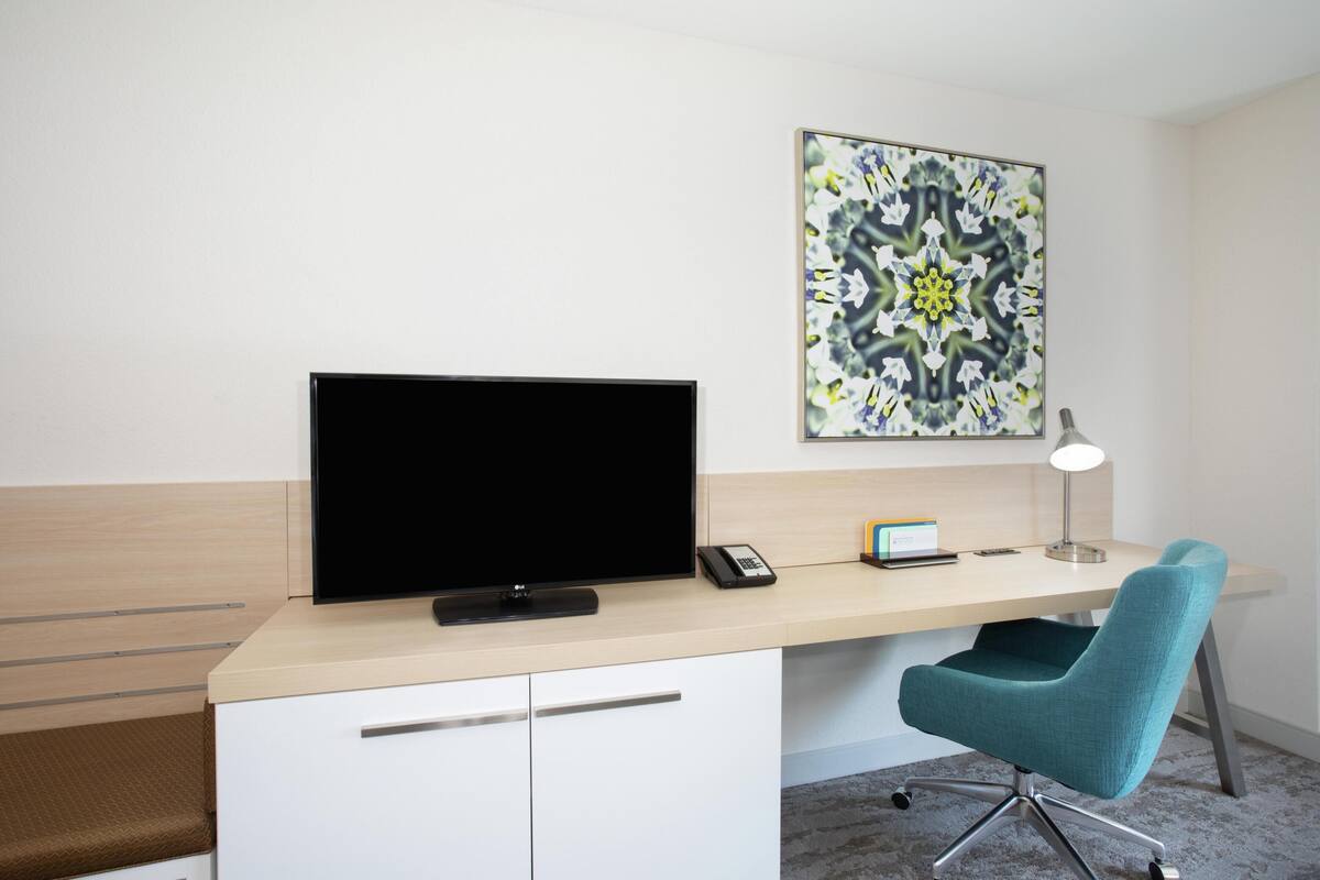 Una habitación de hotel moderna con una televisión de pantalla plana, un escritorio con lámpara, una silla y un elemento decorativo en la pared.