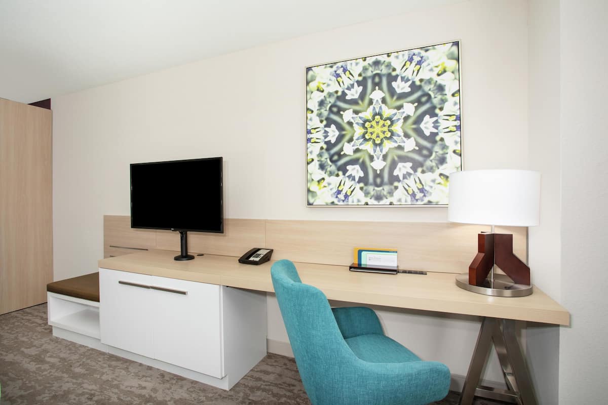 Habitación de hotel con escritorio, televisión de pantalla plana, lámpara, silla, mesita y una obra de arte enmarcada en la pared.