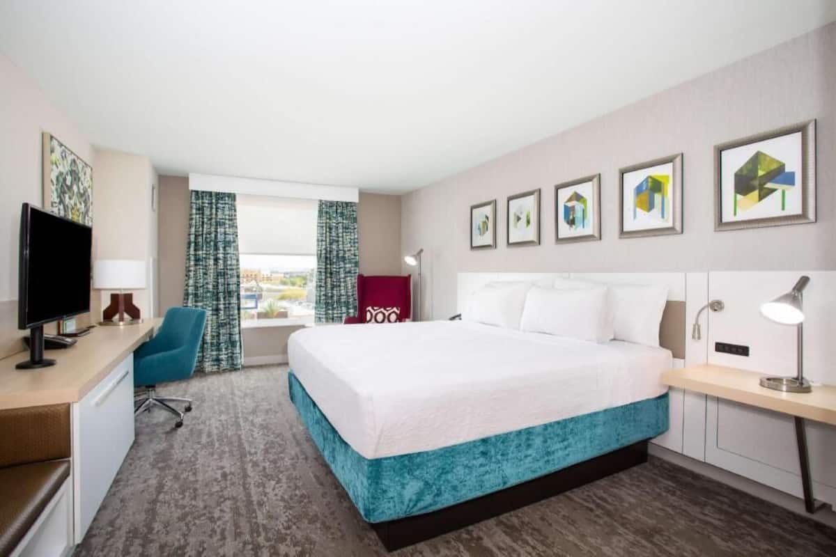 Una habitación de hotel con cama, escritorio, silla, televisión, ventana con cortinas y una obra de arte en la pared.