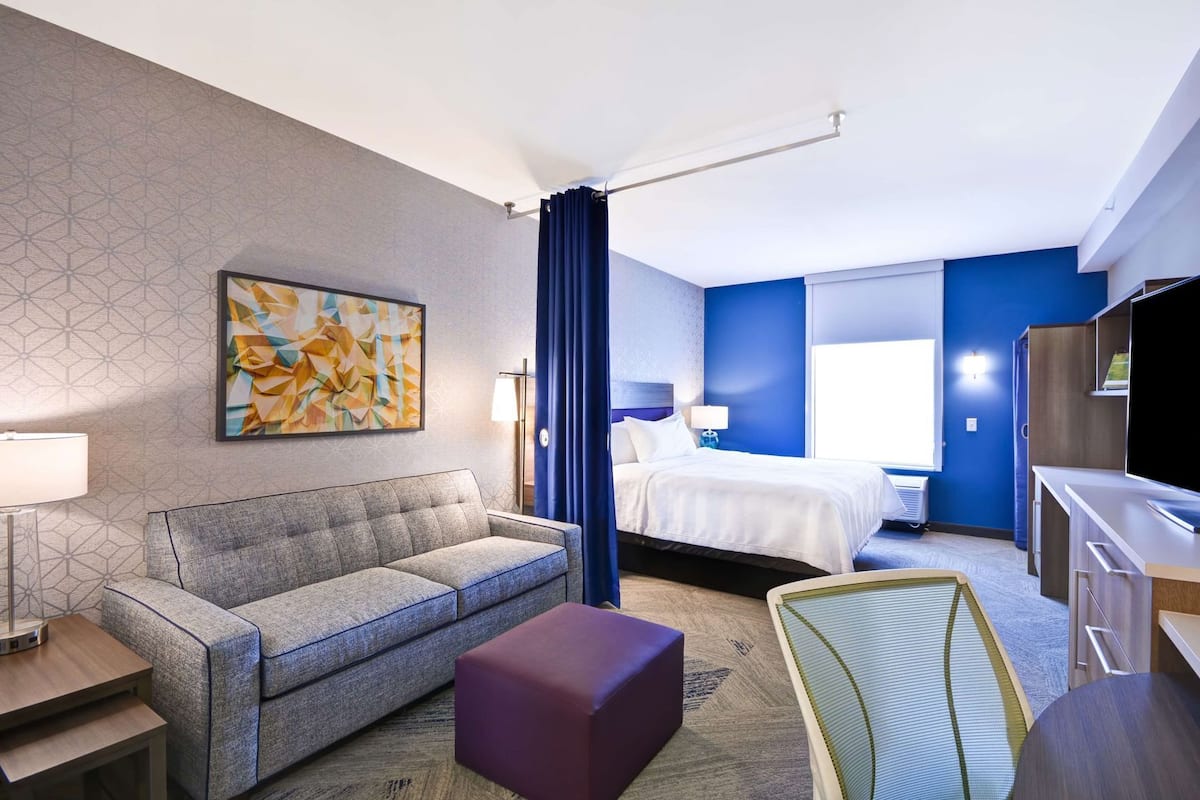 A hotel room with a bed, a sofa, a desk, a TV, and a painting on the wall.