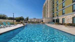 Una piscina rectangular con agua azul cristalina, rodeada de sillas de descanso y un edificio de piedra con grandes ventanales.