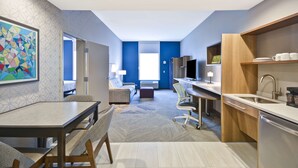 Una habitación de hotel moderna con zona de comedor, kitchenette y un amplio ventanal que proporciona luz natural.
