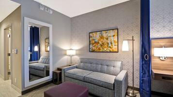 A modern hotel room with a grey sofa, a purple ottoman, a large mirror, a floor lamp, and a wall-mounted artwork.
