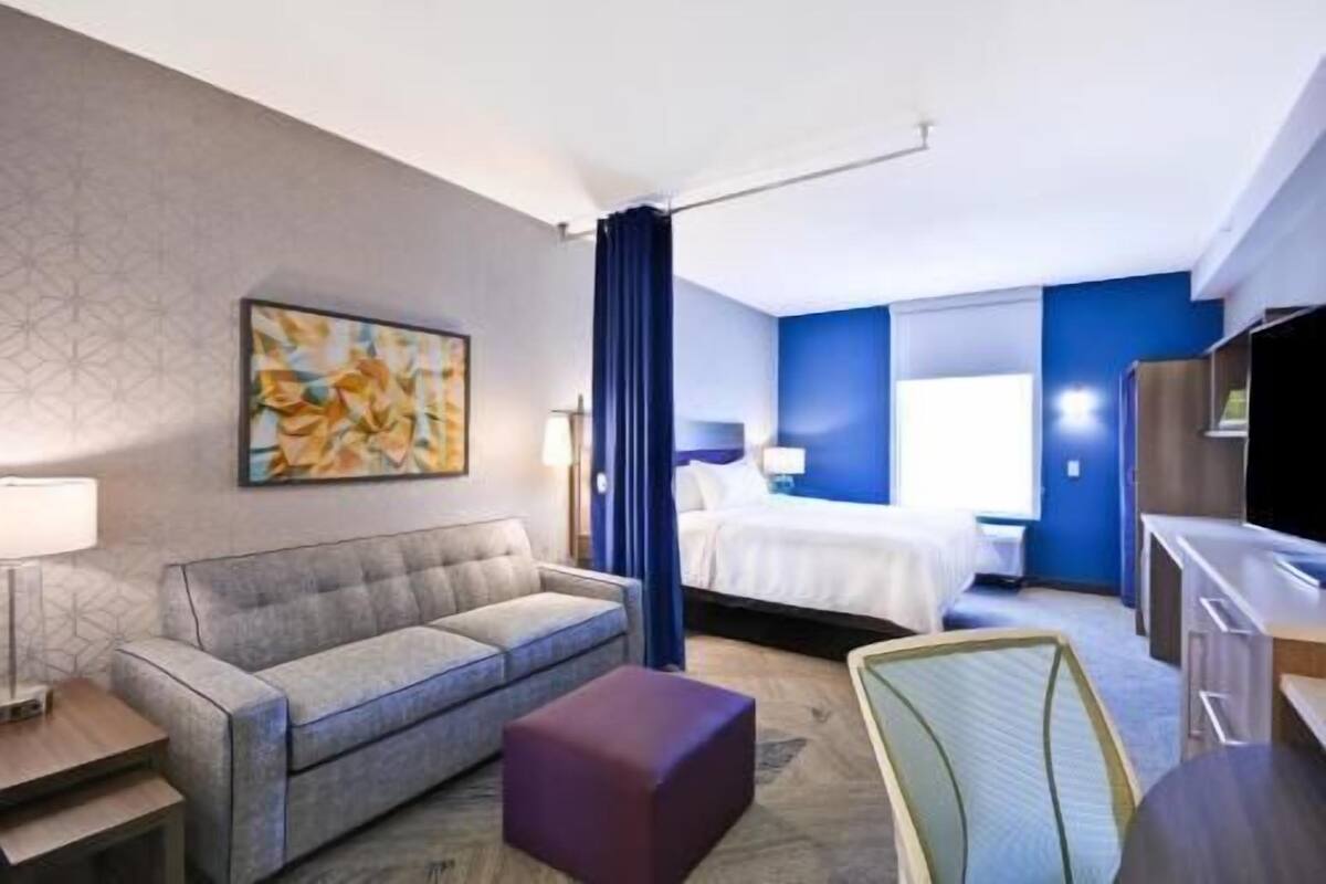 A hotel room with a bed, a sofa, a desk, a chair, and a painting on the wall.