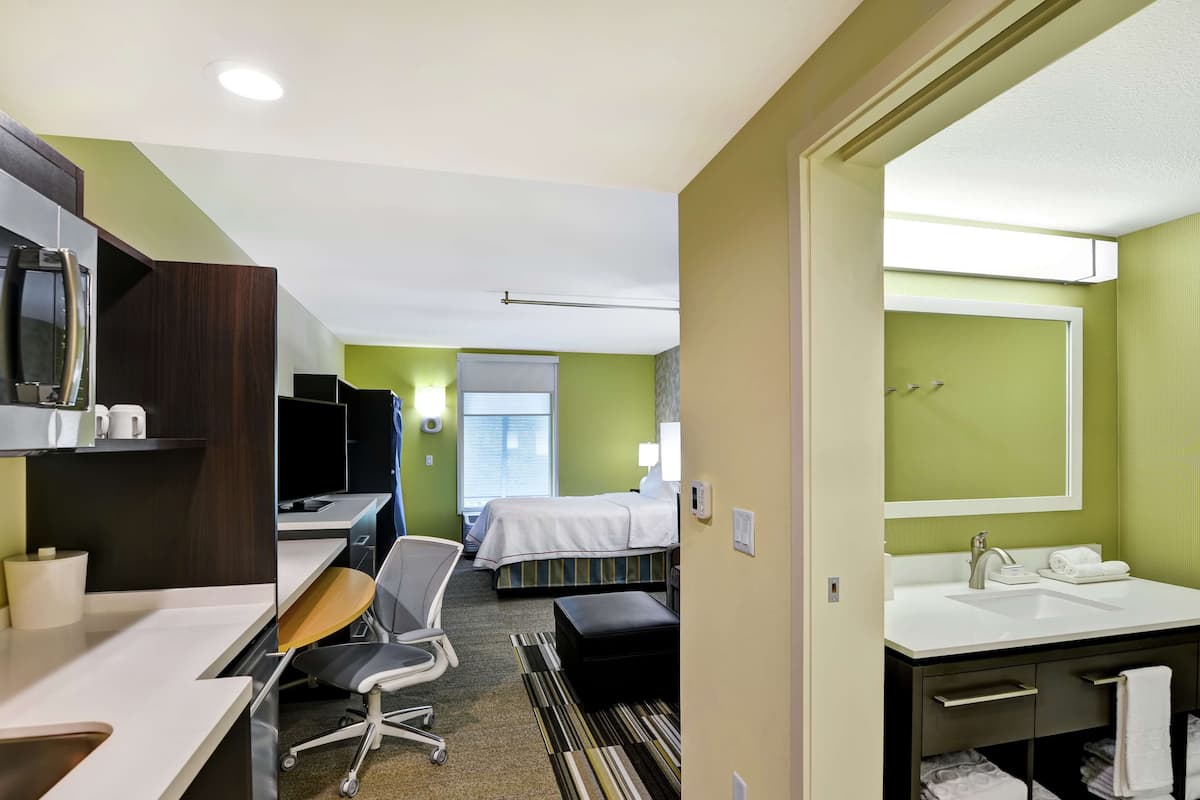 A hotel room with a desk, chair, bed, and a bathroom with a sink and mirror.