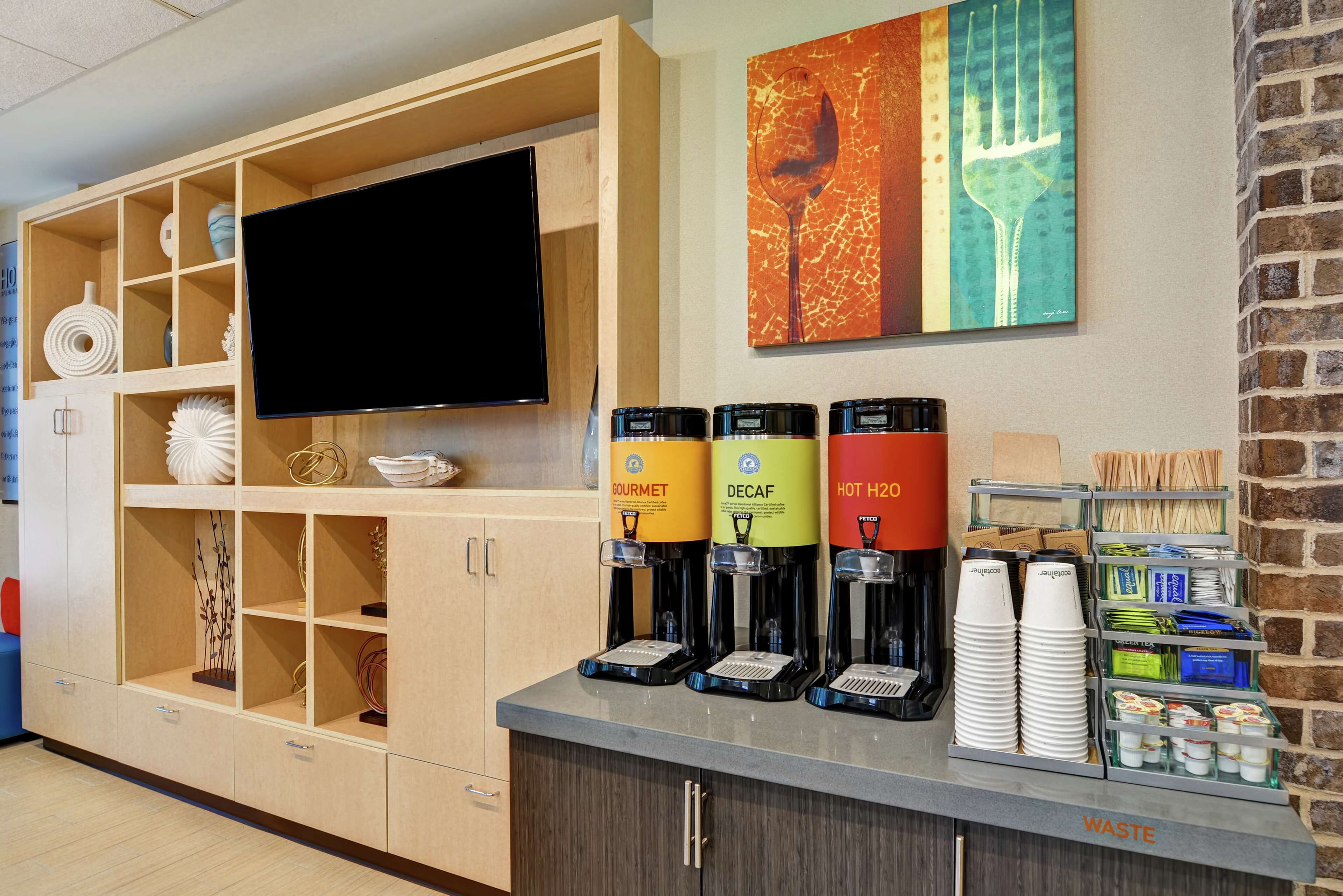 Et hotellets reception område med et fladskærms-tv, drikkevareautomater mærket 'Gourmet', 'Dekaffe' og 'Varm H2O', samt en skraldespand.