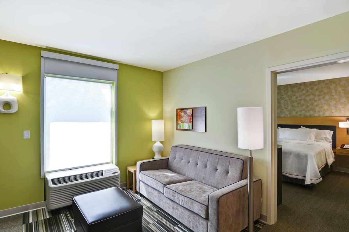 A hotel room with a sofa, a bedside table with a lamp, a bed with white bedding, and a window with blinds.