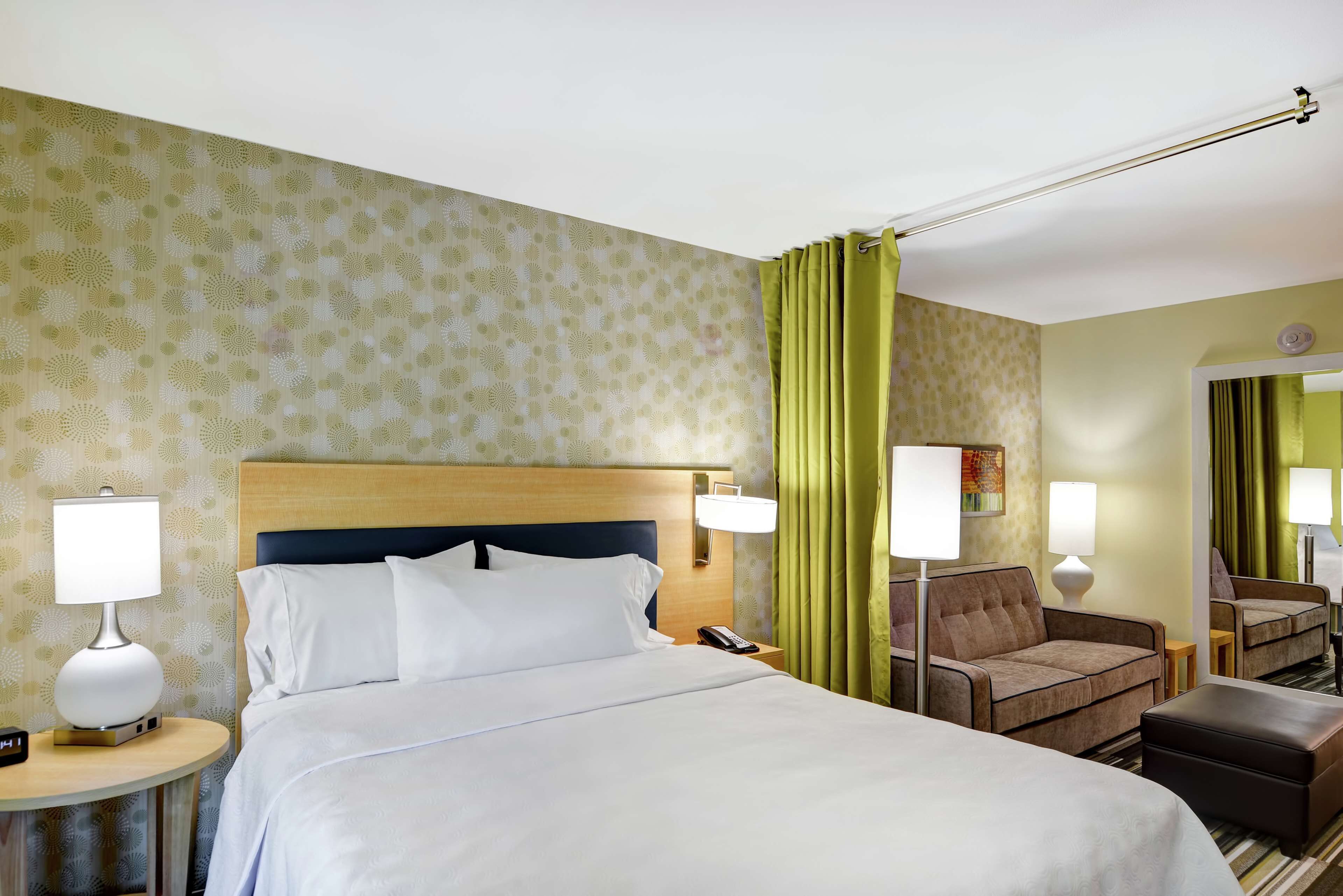 Una habitación de hotel con una cama, dos mesitas de noche con lámparas, una silla, un sofá y un espejo.