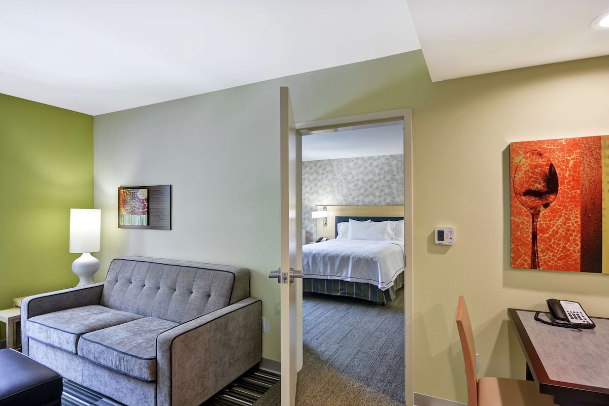 A hotel room with a grey sofa, a bedside table with a lamp, a bed with white bedding, a painting on the wall, and a telephone on a desk.