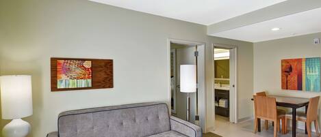 A modern hotel room with a grey sofa, a dining table with chairs, and artwork on the walls.