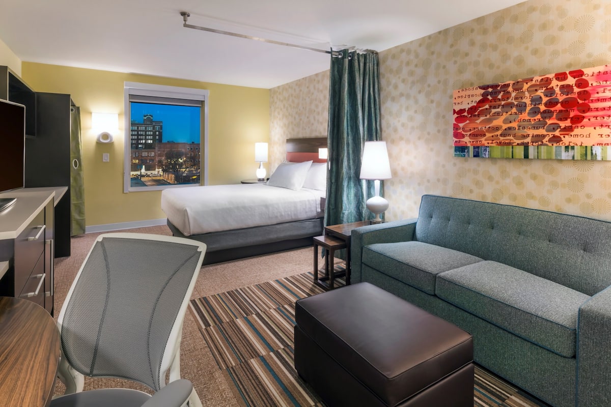 Ein Hotelzimmer mit Bett, einer Sitzecke, einem Sessel, einem Schreibtisch und einem Fenster mit Blick auf die Stadt.