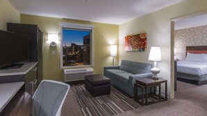 A hotel room with a bed, a sofa, a desk with a TV, a window with a city view, and a lamp. - Home2 Suites by Hilton Kansas City Downtown (Kansas City)