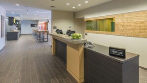 A hotel reception area with a counter, a seating area, and a kitchen in the background. - Home2 Suites by Hilton Kansas City Downtown (Kansas City)