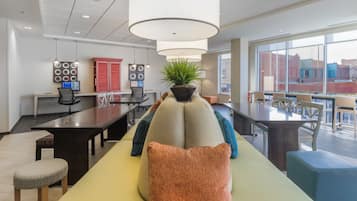 A modern dining area with a long table, cushioned seating, pendant lighting, and a view of buildings through large windows.