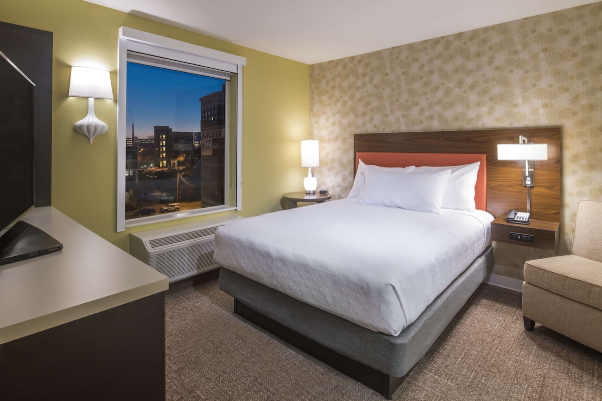 Ein Hotelzimmer mit einem großen Bett, zwei Nachttischen, einem Schreibtisch und einem Fenster mit Blick auf die Stadt.