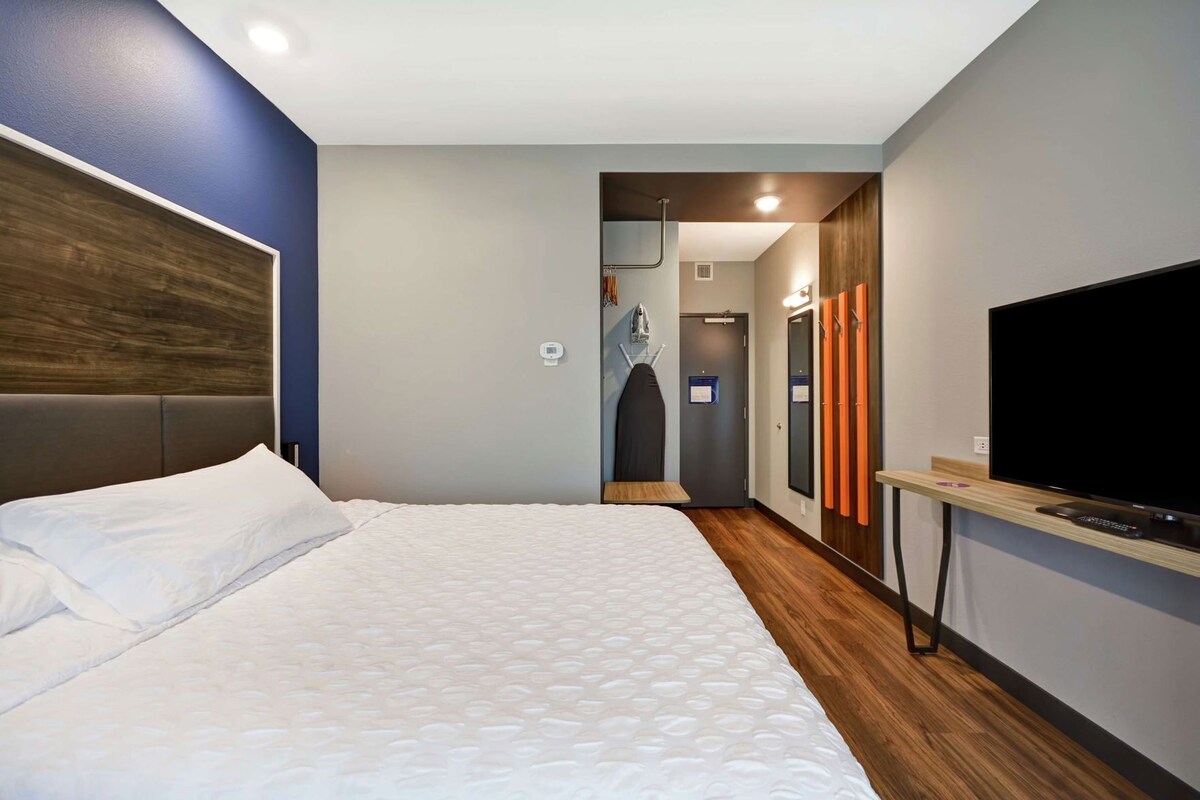 A hotel room with a bed, a TV, a wooden floor, a mirror, and a door leading to a bathroom.