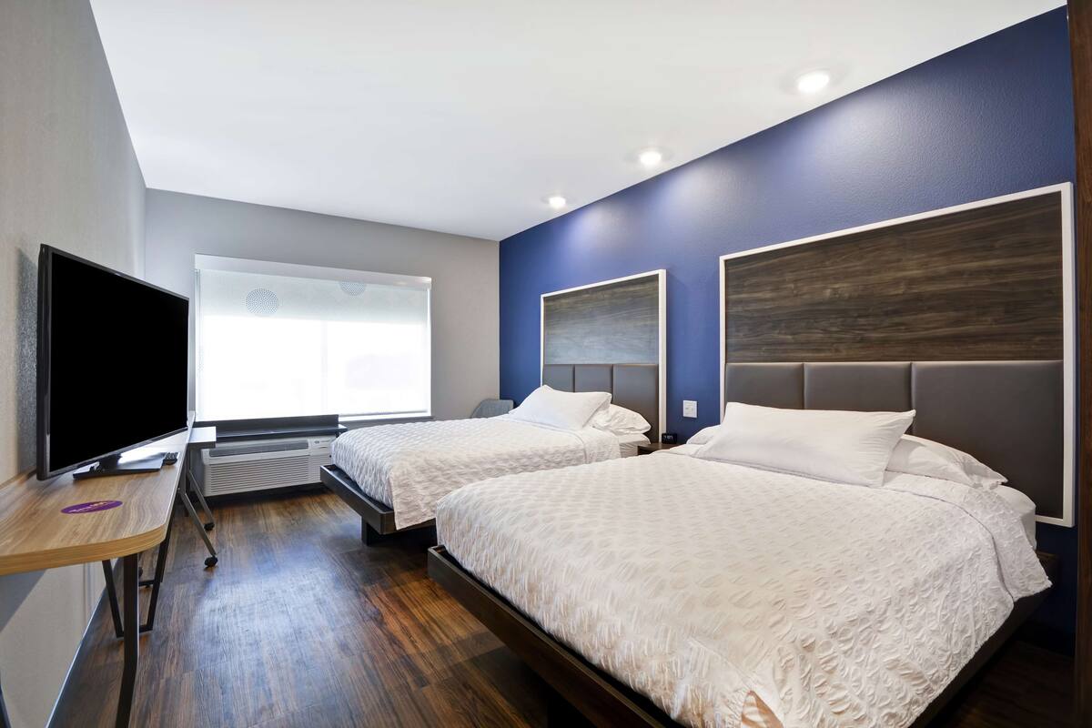 A modern hotel room with two beds, a flat-screen TV, a desk, and a large window.