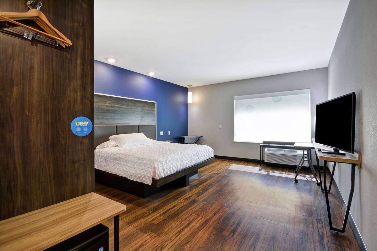 A modern hotel room with a wooden floor, a bed with a white bedspread, a mounted TV, and a large window with blinds.