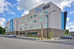 Un hotel de diseño moderno, con un llamativo letrero 'tru' y la marca 'Hilton'. Tiene varios pisos con diferentes tipos de ventanas y un área de estacionamiento al frente.