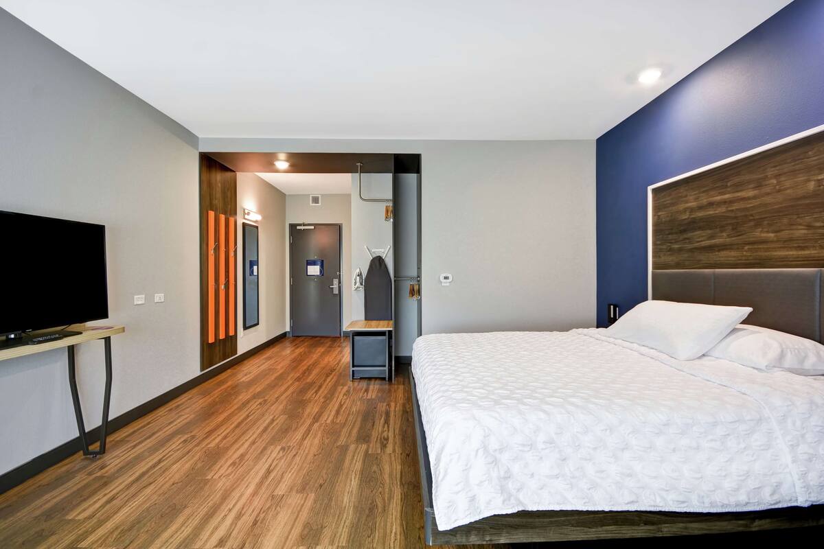 A modern hotel room with a large bed, a flat-screen TV, a wooden floor, and a view of the hallway.