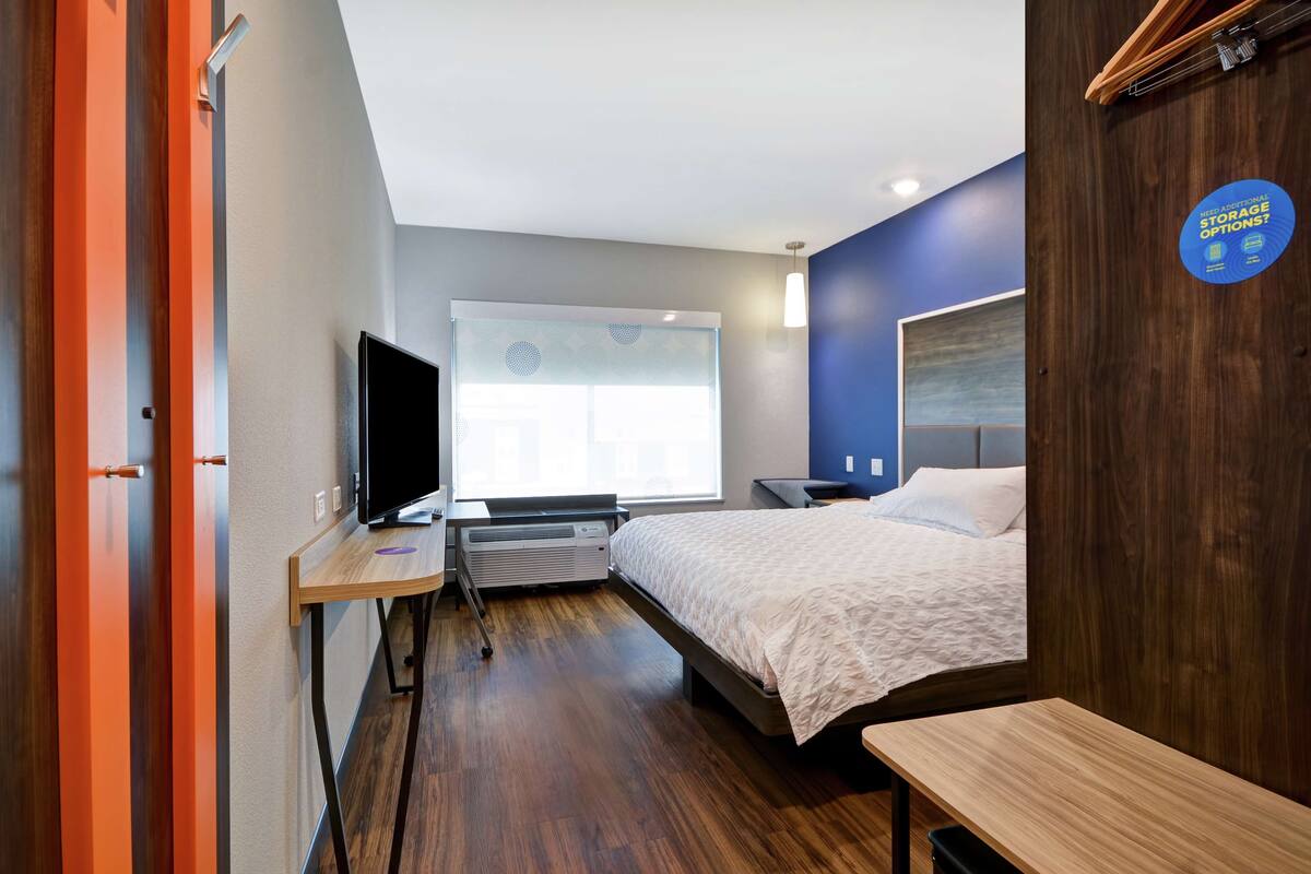 A modern hotel room with a bed, a desk with a computer, a television, and a window with blinds.