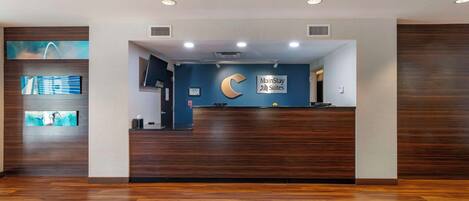 Un'area reception con una scrivania in legno scuro, una parete blu con il logo MainStay Suites e un'ampia finestra che mostra una vista sulla città.