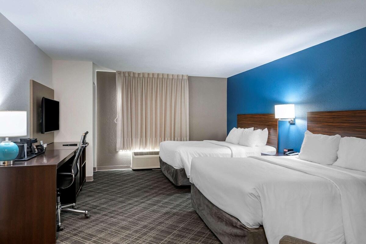 Habitación de hotel con dos camas, un escritorio con computadora, televisor y ventana con cortinas.