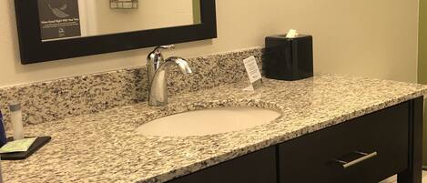 A bathroom with a granite countertop, a sink, a mirror, a towel rack, and a black soap dispenser.