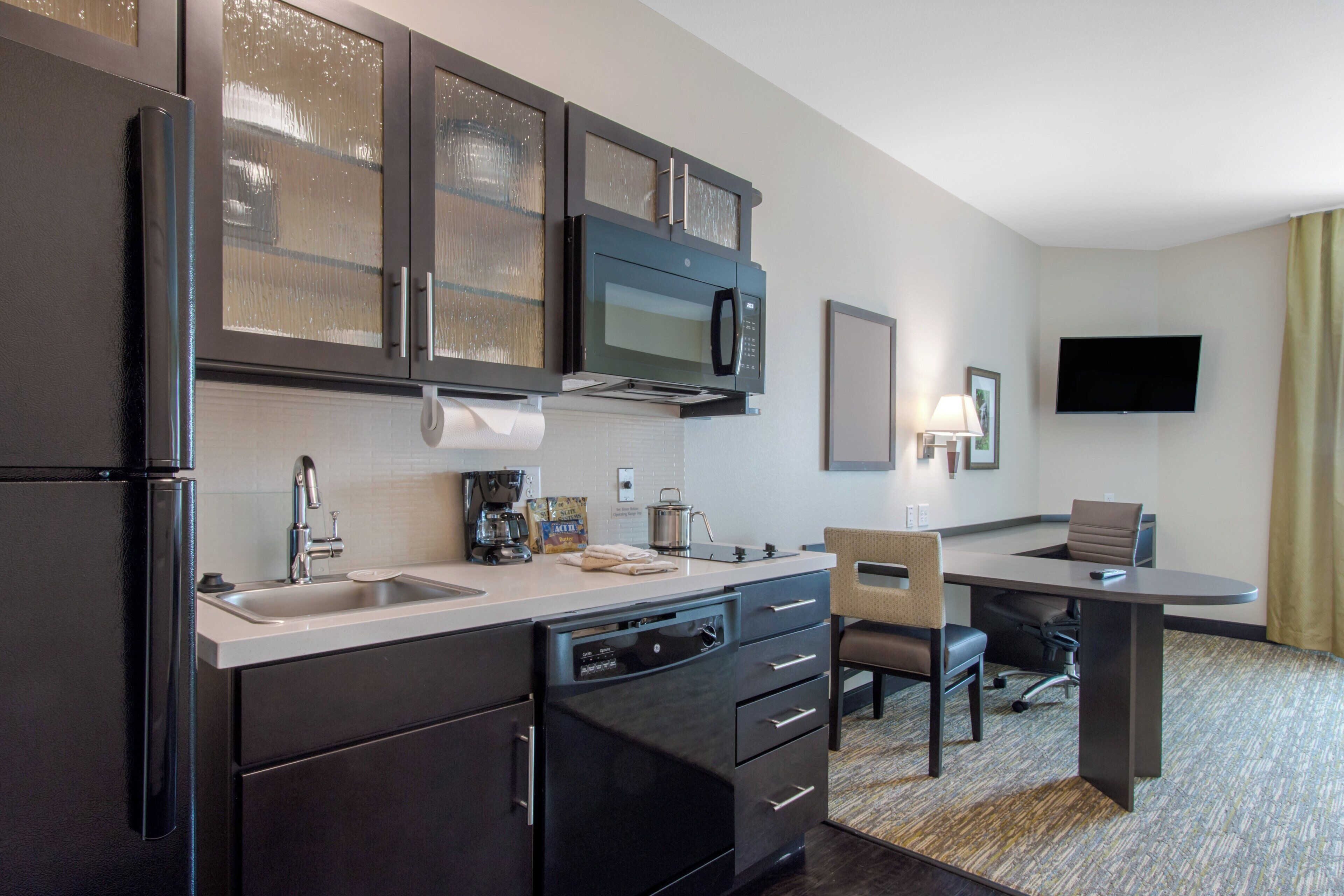 A compact hotel kitchen with a refrigerator, microwave, sink, and a small dining area with a chair and table.