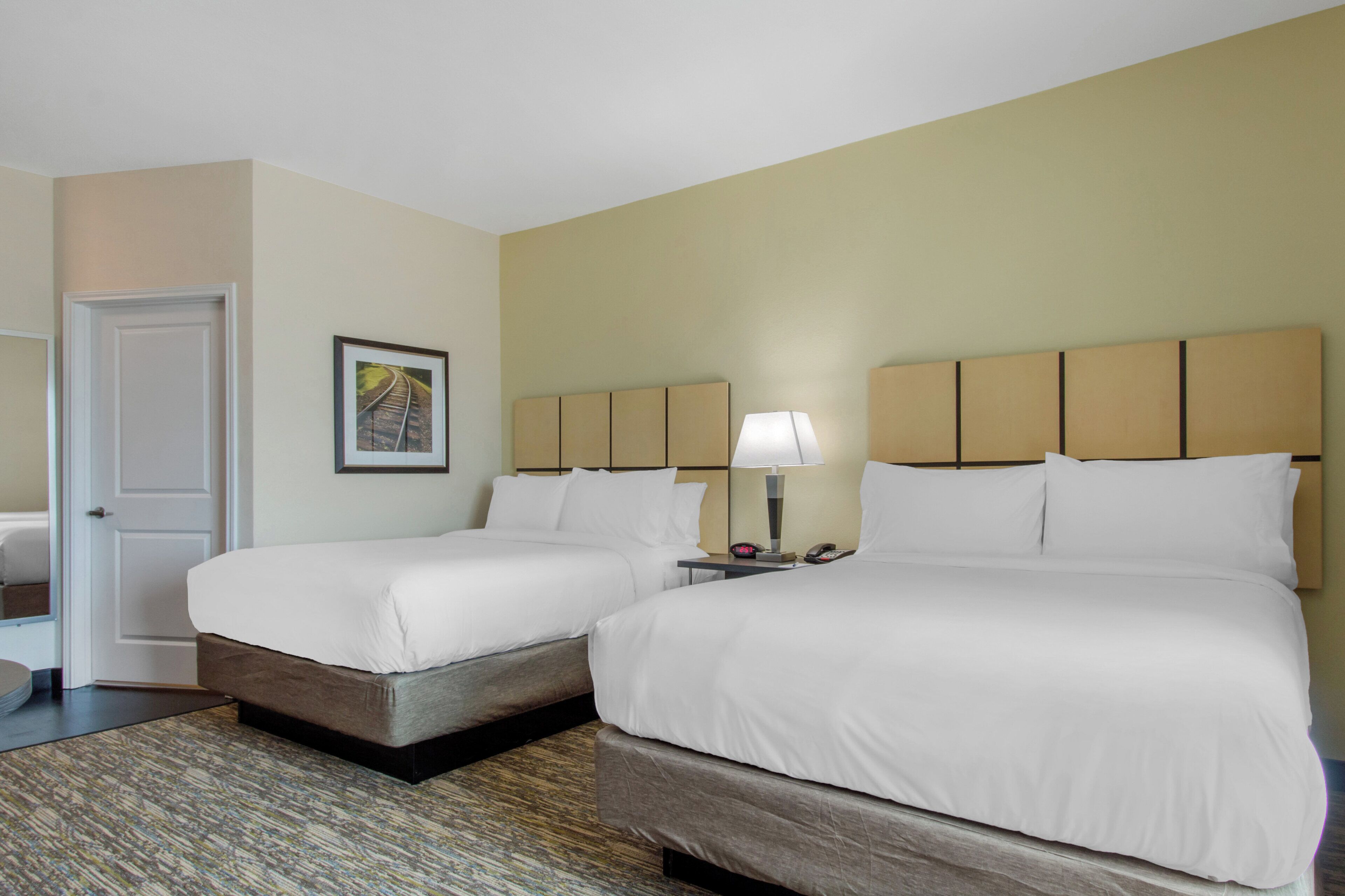 A hotel room with two beds, a nightstand with a lamp, a framed picture on the wall, and a door.