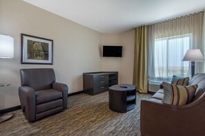 A hotel room with a brown leather armchair, a small round table, a flat-screen TV mounted on the wall, a framed picture, and a window with curtains. - Candlewood Suites San Antonio Lackland AFB Area by IHG (San Antonio)