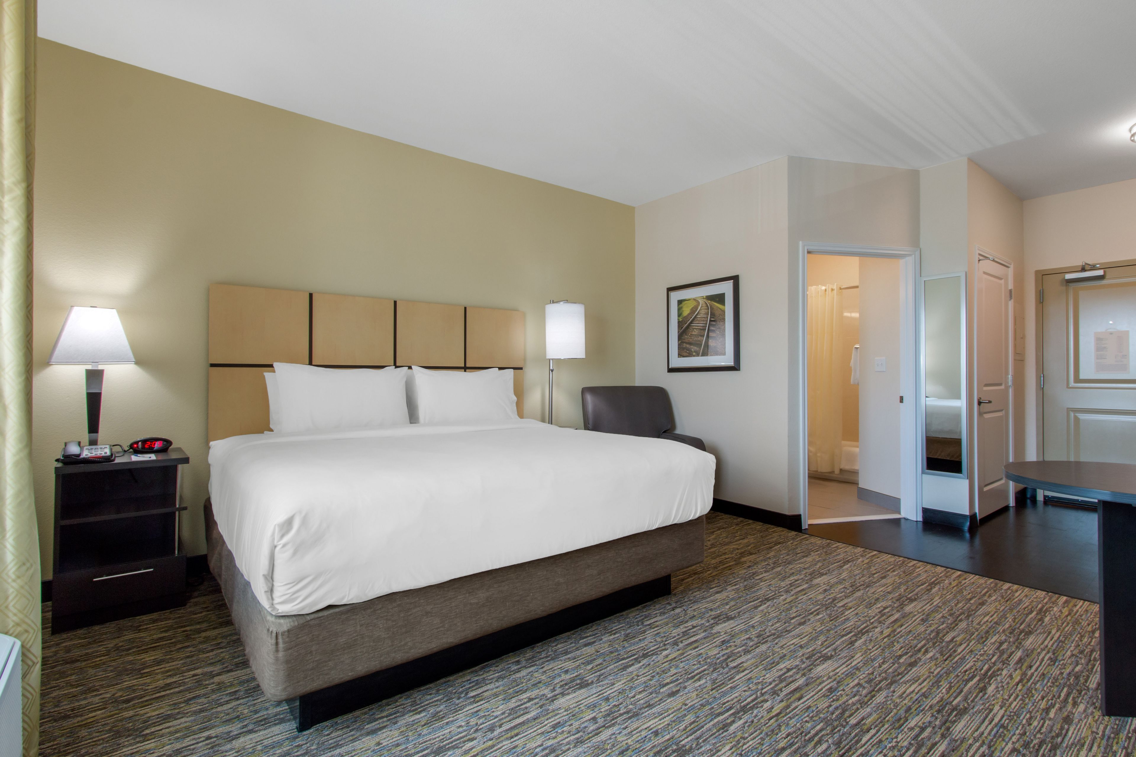 A hotel room with a large bed, a desk, and a chair. There is a lamp on the desk and a picture on the wall.