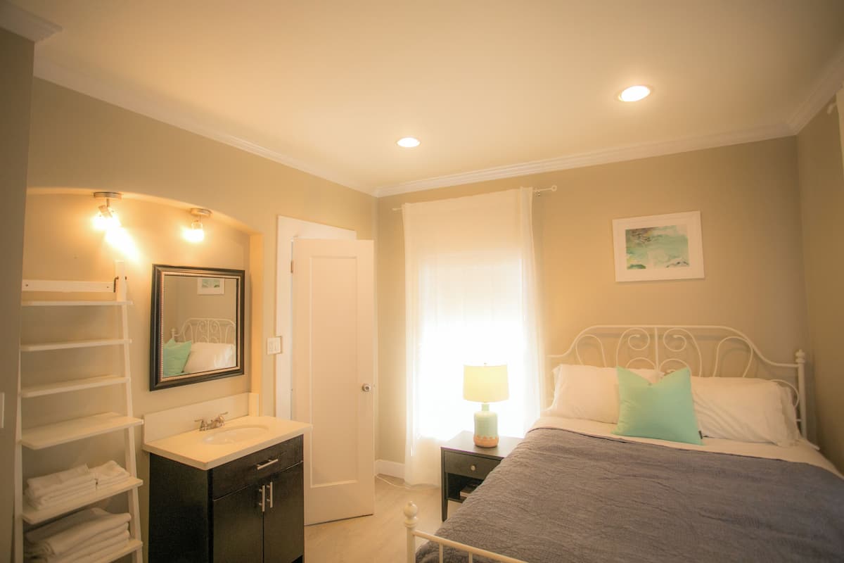 Une chambre à coucher avec un lit, une table de chevet avec une lampe, une commode, un miroir et une fenêtre avec des rideaux.