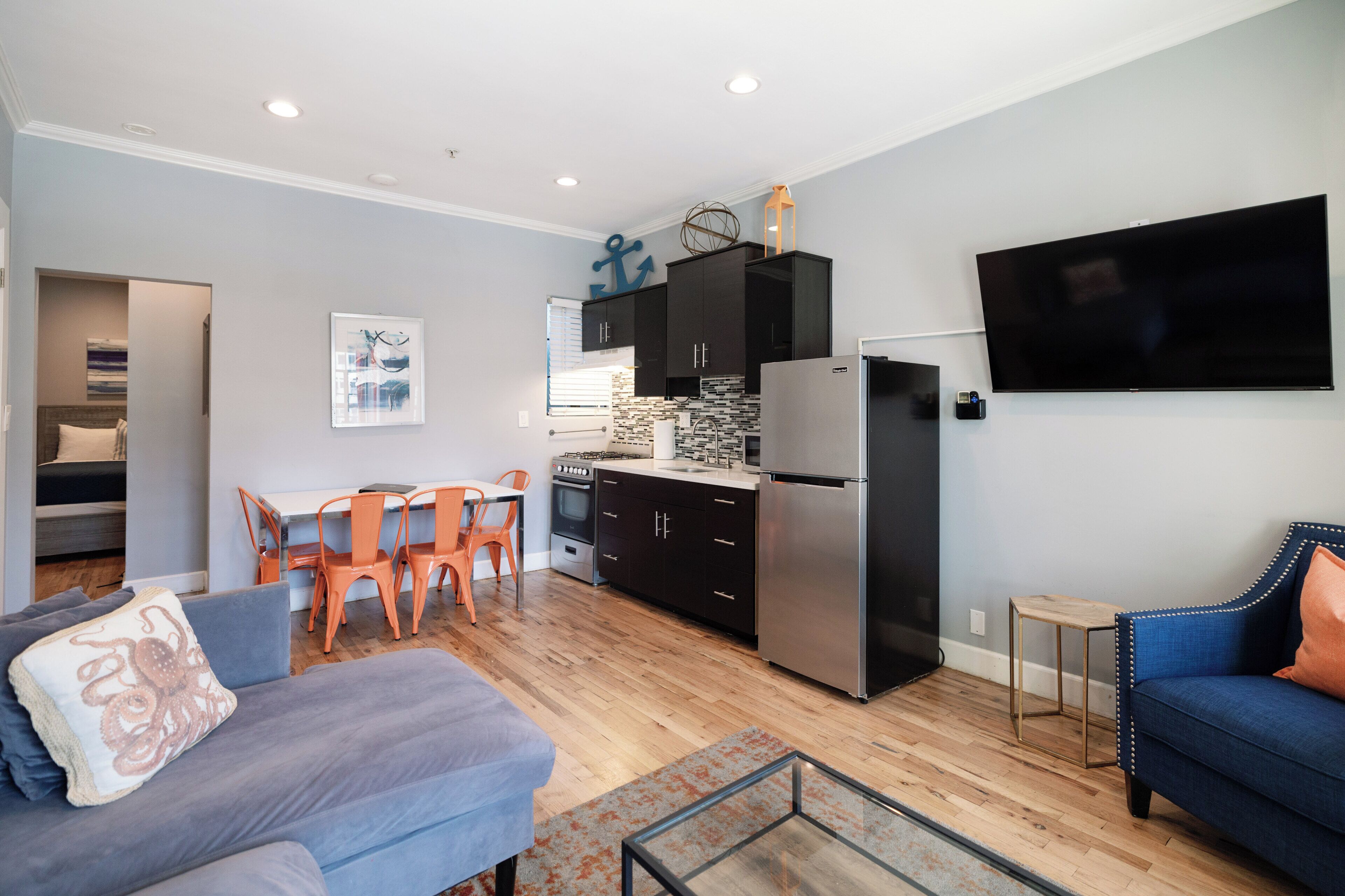 A modern living room with a grey sofa, a blue sofa, and a glass coffee table. There is a kitchen area with black cabinets, a stainless steel refrigerator, and a flat-screen TV mounted on the wall.