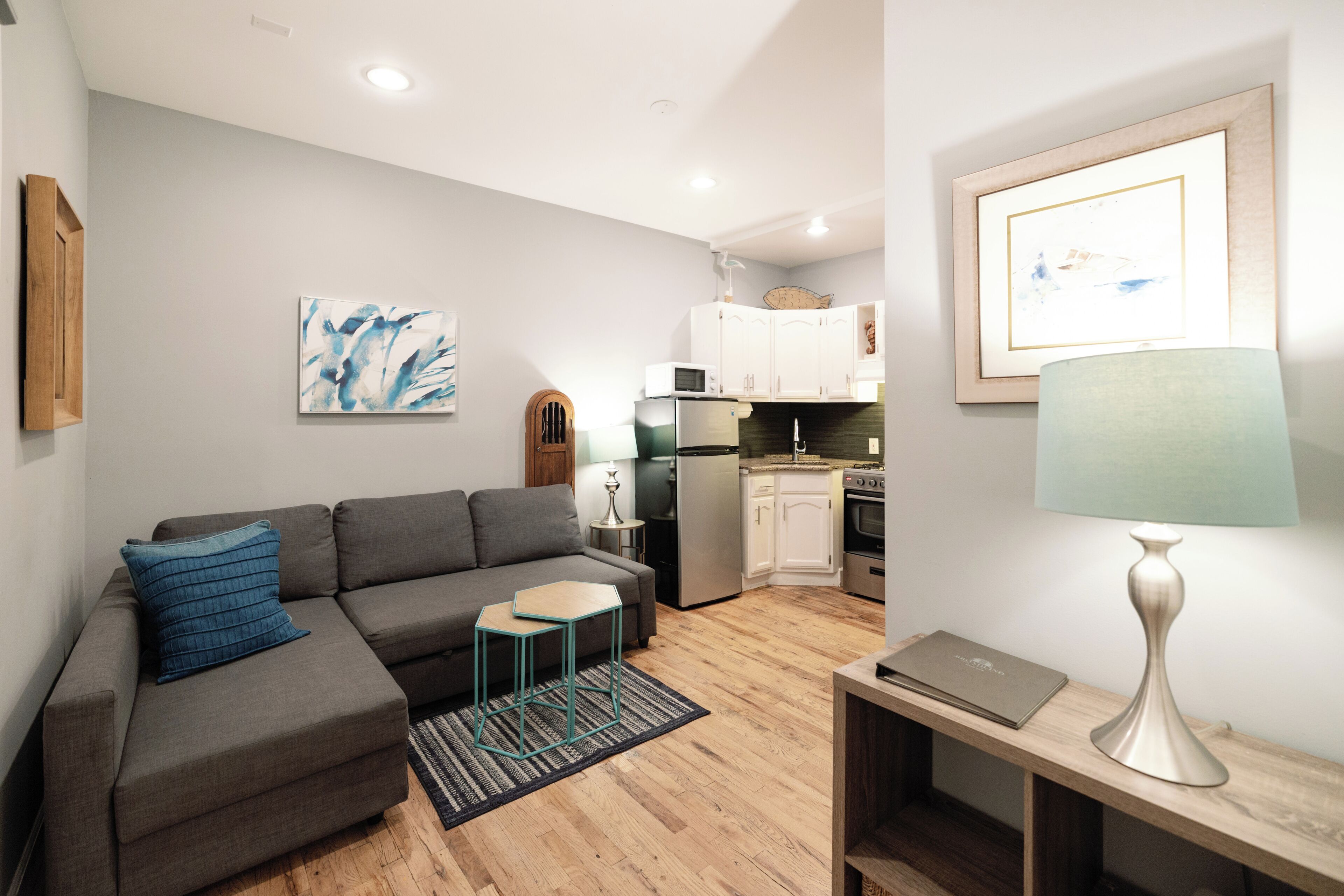 A modern living room with a grey sectional sofa, a small teal stool, and a wooden coffee table. A framed picture hangs on the wall, and a lamp is on a side table.